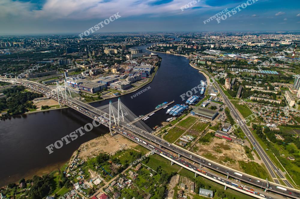 Панорамный вид с дрона на Санкт-Петербург с современным вантовым мостом через реку Неву. Масштабная транспортная инфраструктура, городские магистрали и архитектура северной столицы России создают впечатляющий урбанистический пейзаж. Аэрофотосъёмка подчёркивает красоту инженерных конструкций, динамику мегаполиса и живописные виды Невы. Фотография идеально подходит для публикаций о современном Санкт-Петербурге, мостах России, городской архитектуре, транспортной инфраструктуре и путешествиях по России.